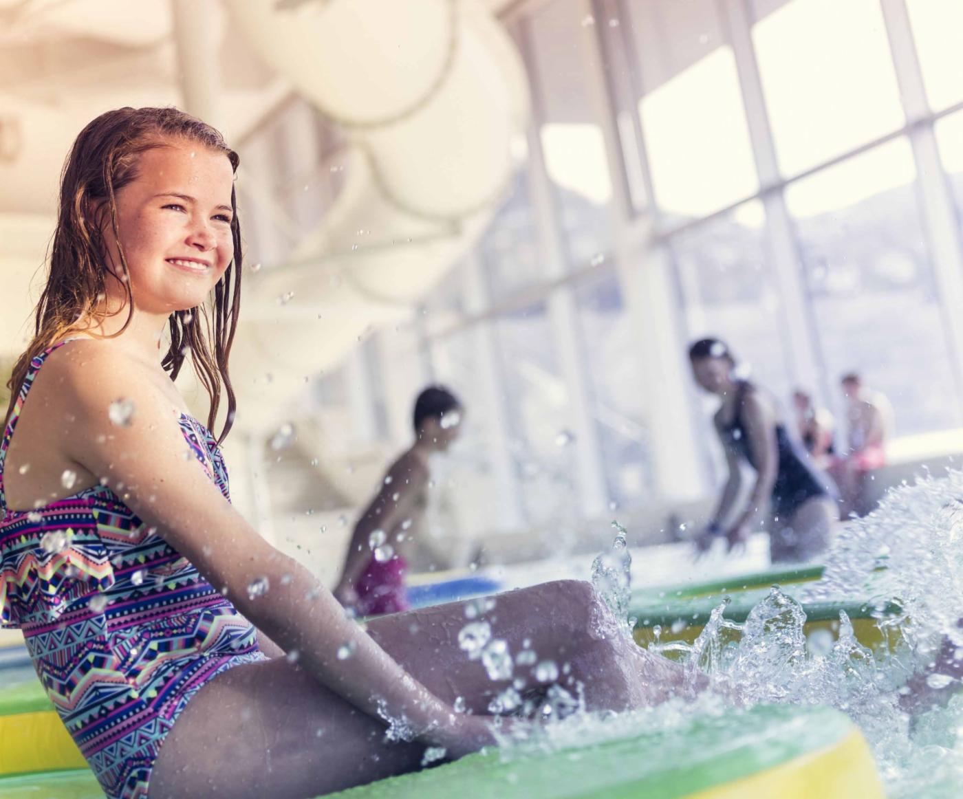 En jente smiler mens hun sitter ved et innendørs svømmebasseng. 