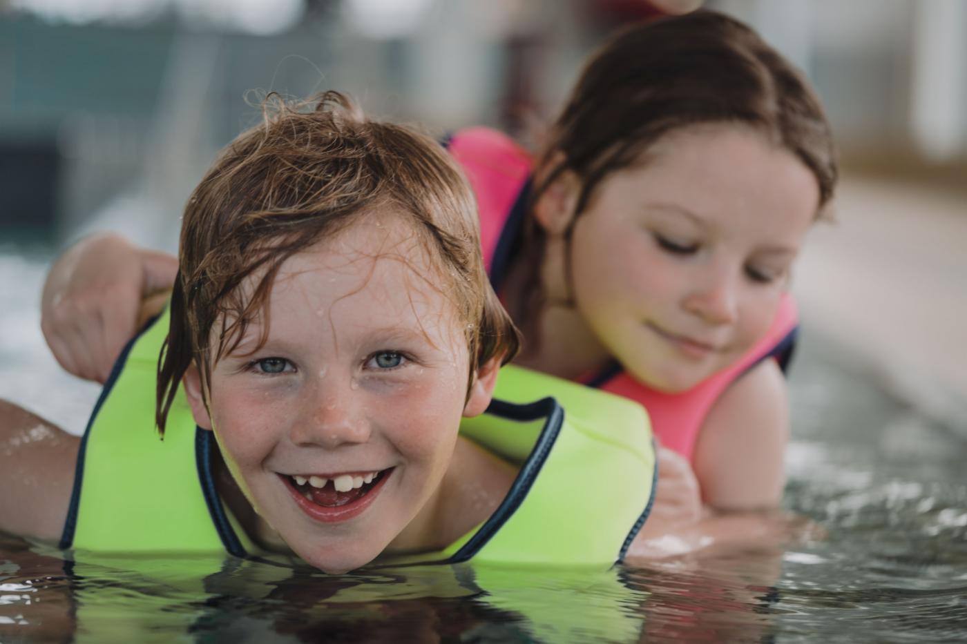 En gutt smiler til kamera mens han og en jente er i et innendørs svømmebasseng. 
