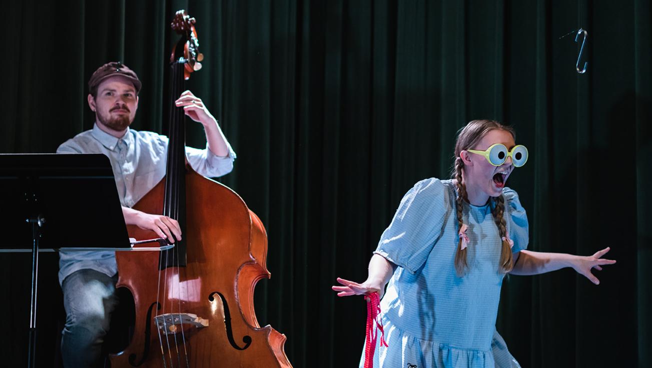 En mann spiller kontrabass mens en dame ikledd morsomme briller slår ut med armene og synger. 