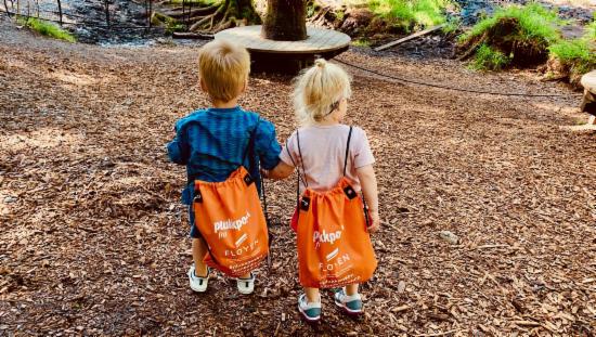 To små barn går i skogen med ryggen mot kamera. På ryggen har den hver sin oransje sekk.